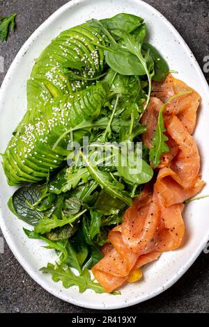 Bassa di un glucide insalata. Gli spinaci, rucola con insalata di avocado e salmone. Nero lo sfondo di calcestruzzo, piastra bianca, vista dall'alto Foto Stock