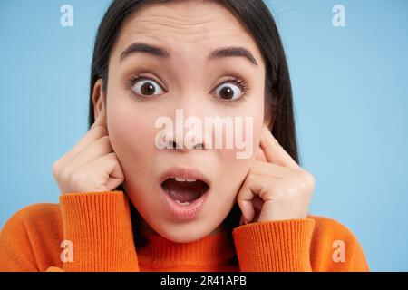 Primo piano ritratto di ragazza asiatica chiude le orecchie da forte rumore, sembra scioccato alla macchina fotografica, si trova su sfondo blu Foto Stock