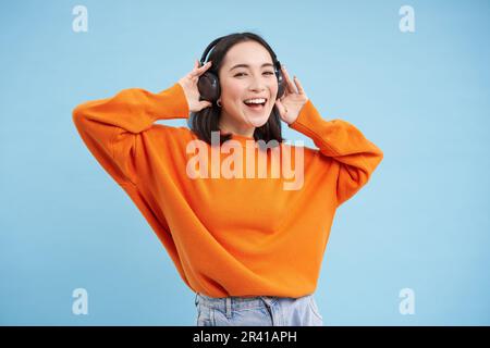 Felice donna cinese in cuffie, ascolta la musica, ama la canzone preferita nella sua playlist, si trova su sfondo blu Foto Stock