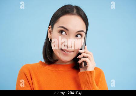 Allegra ragazza asiatica parla sul telefono cellulare, parla con qualcuno al telefono, si trova su sfondo blu Foto Stock