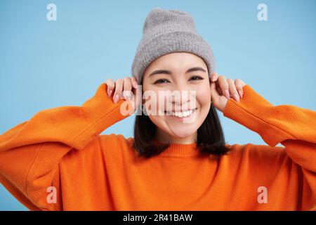 Primo piano ritratto di felice, elegante ragazza coreana mette su caldo cappello, sorride e sembra gioioso, si trova contro sfondo blu studio Foto Stock