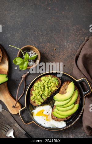 Pane integrale toast avocado a fette e uovo in camicia su fondo di pietra. Concetto di colazione sana, sandwich con avocado e. Foto Stock