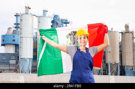 Ragazza felice in abiti da lavoro e elmetto con bandiera del messico in piedi di fronte a scenario industriale Foto Stock