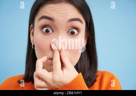 Primo piano ritratto di donna giapponese chiude il naso da cattivo odore, si trova su sfondo blu Foto Stock