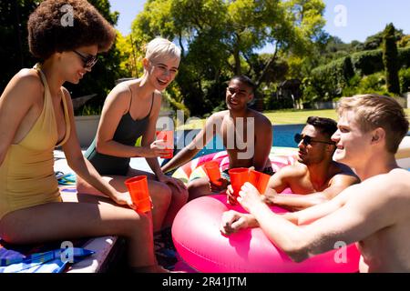 Felice gruppo variegato di amici che hanno partito in piscina, facendo brindisi in piscina in giardino Foto Stock