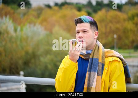 Un giovane uomo che indossa un impermeabile giallo che registra un messaggio audio sullo smartphone per strada in autunno. Foto Stock