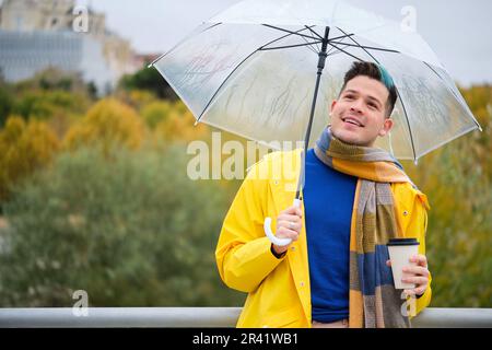 Giovane uomo che indossa un impermeabile giallo con un caffè e un ombrello in strada in autunno. Foto Stock