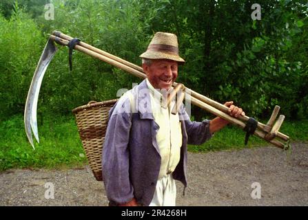 Maramures County, Romania, circa 2001. Uomo locale sulla strada di campagna che porta due falci. Foto Stock