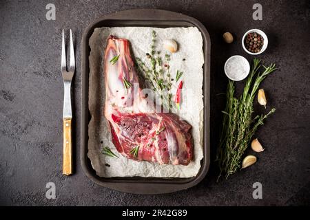 Crudo fresco di carne di agnello in teglia da forno, erbe e la forcella sulla pietra nera dello sfondo. Foto Stock