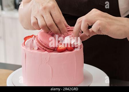 Donna pasticceria chef decorare torta rosa con macaroon e bacche, primo piano. Processo di fabbricazione della torta, fuoco selettivo Foto Stock