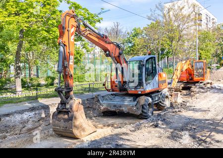 Gli escavatori gommati e cingolati di grandi dimensioni sono parcheggiati in una trincea scavata nel mezzo di una strada cittadina in una giornata di sole primavera. Foto Stock