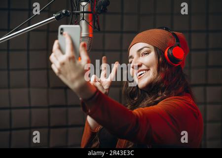 Bella donna attraente blogger utilizzando lo smartphone per selfie e registrazione video vlog per i suoi abbonati al suono studio di registrazione vicino a un profissio Foto Stock