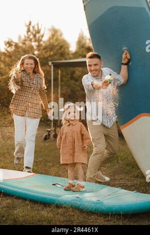 la famiglia riposa accanto alla casa mobile. Papà, mamma e figlia giocano sulle tavole di sup con pistole ad acqua vicino al camper. Foto Stock