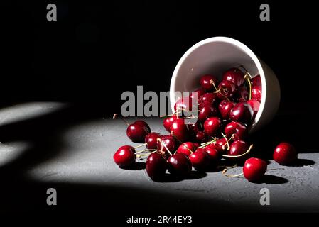 Ciotola carica di ciliegie rosse su fondo scuro. Tazza rovesciata che sparge ciliegie ricche e sane Foto Stock