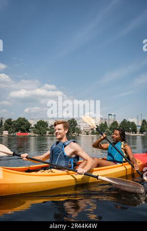 coppia multietnica attiva e positiva nei giubbotti di vita che trascorrono il fine settimana ricreativo navigando in kayak sportivo sul fiume della città sotto il cielo blu con bianco Foto Stock