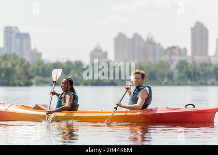 incantevole donna afroamericana e sportivo uomo rosso in giubbotti di vita guardando la macchina fotografica durante la navigazione in kayak lungo il fiume con sfocata città sc Foto Stock