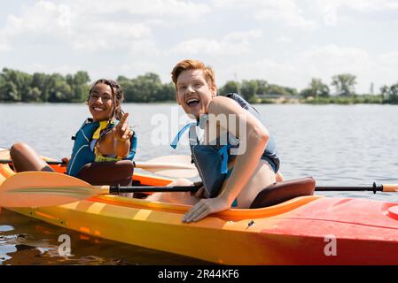 eccitato uomo rosso con bocca aperta guardando la macchina fotografica vicino allegra afroamericana donna in giubbotto di vita che punta con il dito mentre naviga in kayak Foto Stock