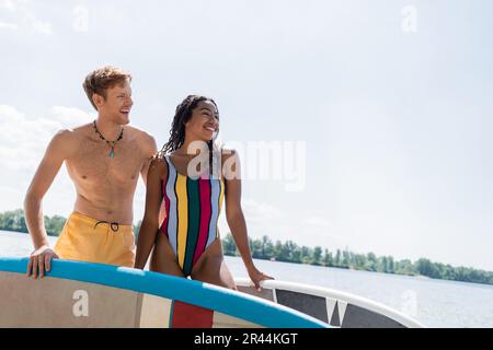 donna afro-americana spensierata e carina in costume da bagno a righe e giovane uomo rosso in pantaloncini gialli sorridenti e guardando lontano da tavole di sup ou Foto Stock