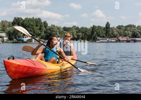 donna afro-americana e giovane uomo rosso overjoyed in giubbotti di vita trascorrere il fine settimana estivo in barca a vela in kayak con pagaie sul lago con scena verde Foto Stock