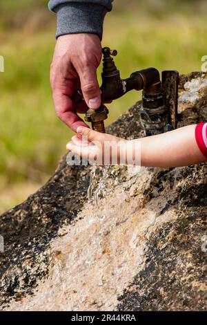 Il bambino pulisce le mani da un rubinetto esterno che esce da una pietra, spruzzi rinfrescanti senza sprecare acqua Foto Stock