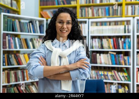 Ritratto di giovane insegnante all'interno della biblioteca accademica didattica, donna con braccia incrociate sorridente e guardando la macchina fotografica, ricercatore insegnante latino-americano di successo. Foto Stock