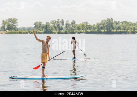 allegro uomo rosso guardando via e indicando con mano vicino sportivo afro-americano donna in costume da bagno colorato vela su tavola di sup il giorno d'estate Foto Stock