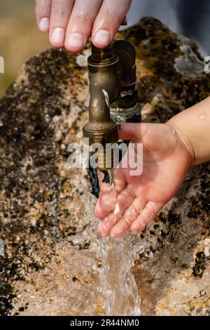 Il bambino pulisce le mani da un rubinetto esterno che esce da una pietra, spruzzi rinfrescanti senza sprecare acqua Foto Stock