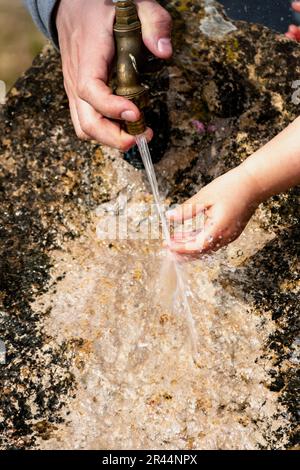 Il bambino pulisce le mani da un rubinetto esterno che esce da una pietra, spruzzi rinfrescanti senza sprecare acqua Foto Stock