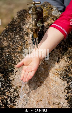 Il bambino pulisce le mani da un rubinetto esterno che esce da una pietra, spruzzi rinfrescanti senza sprecare acqua Foto Stock