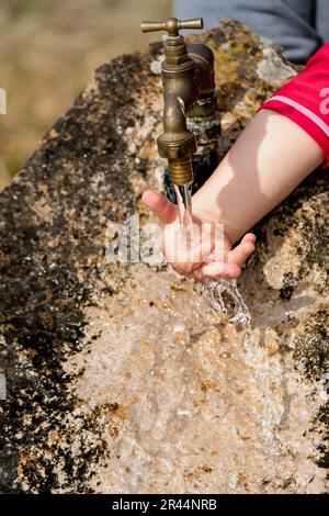 Il bambino pulisce le mani da un rubinetto esterno che esce da una pietra, spruzzi rinfrescanti senza sprecare acqua Foto Stock