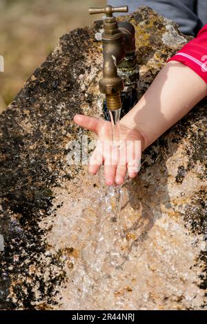 Il bambino pulisce le mani da un rubinetto esterno che esce da una pietra, spruzzi rinfrescanti senza sprecare acqua Foto Stock