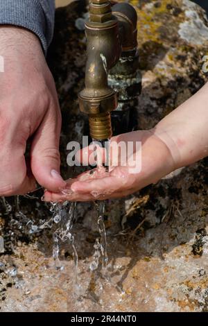 Il bambino pulisce le mani da un rubinetto esterno che esce da una pietra, spruzzi rinfrescanti senza sprecare acqua Foto Stock