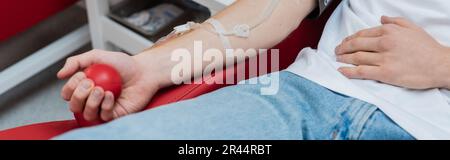 vista ritagliata del volontario con set trasfusion schiacciare palla di gomma mentre si siede su comoda sedia medica nel moderno centro di donazione di sangue, banne Foto Stock