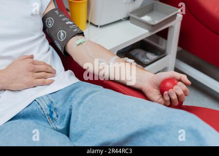vista parziale del donatore con set per trasfusione schiacciare la palla di gomma mentre si siede su una comoda sedia vicino a un vassoio medico sfocato e una tazza di plastica nel sangue Foto Stock