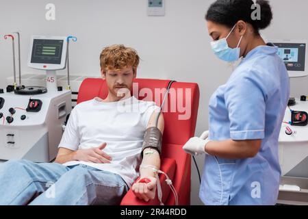 volontario redhead con set trasfusion schiacciare palla di gomma mentre si siede su sedia medica vicino a apparecchiature automatiche e infermiere multirazziale in medicina Foto Stock