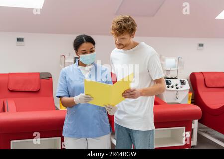 volontario redhead e infermiere multirazziale in uniforme, maschera medica e guanti in lattice guardando la cartella di carta vicino a sedie mediche e macchine per trasfusione Foto Stock