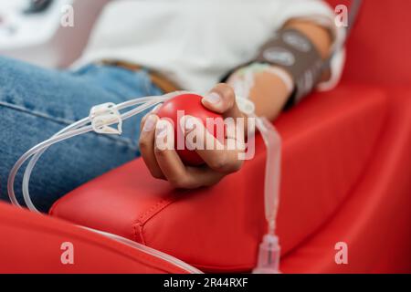 fuoco selettivo della sfera di gomma in mano di donna multirazziale con set trasfusionale seduto su comoda sedia medica e donando sangue in laboratorio Foto Stock