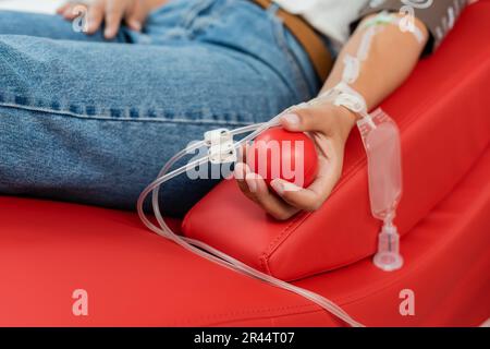 vista parziale della donna multirazziale con set trasfusionale che tiene la palla di gomma mentre si siede su comoda sedia medica durante la donazione di sangue in laboratorio Foto Stock