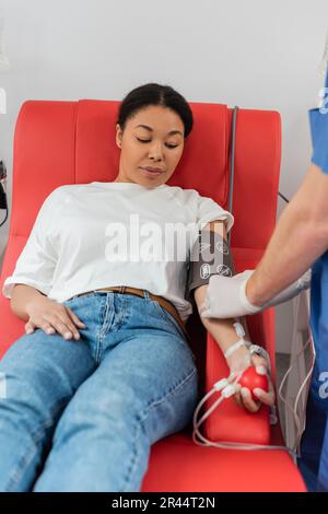 operatore sanitario che collega trasfusione di sangue set a donna multirazziale seduta su comoda sedia medica nel bracciale per la pressione sanguigna e schiacciare la spazzatura Foto Stock