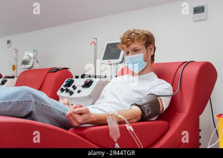 giovane donatore di testa rossa in maschera medica e set per trasfusione schiacciare la palla di gomma mentre si siede su una comoda sedia vicino ad apparecchiature automatiche durante il sangue Foto Stock