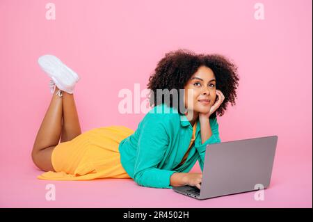 A tutta lunghezza positiva bella afro-americana o brasiliana, vestita elegantemente, sdraiata sul pavimento con un laptop aperto, su sfondo rosa isolato, guardando penosamente lontano, pensando, sognando, sorridendo Foto Stock