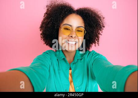 Bella donna alla moda brasiliano o ispanica, blogger, indossare camicia verde casual e occhiali alla moda, scattare foto selfie, guarda la macchina fotografica, sorrisi, stare in piedi su sfondo isolato rosa pastello Foto Stock