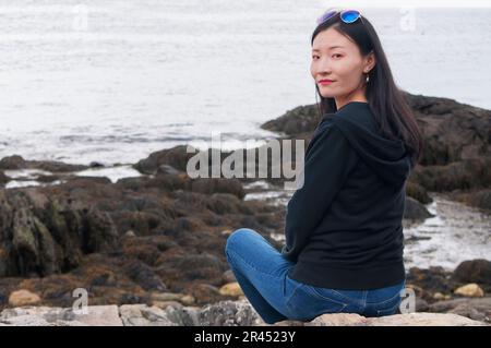 Una donna cinese seduta sulla roccia della costa della baia di casco nel Maine in un giorno coperto. Foto Stock