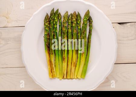 Asparagi alla griglia adagiati su un piatto bianco in ceramica su un tavolo di legno, macro, vista dall'alto. Foto Stock