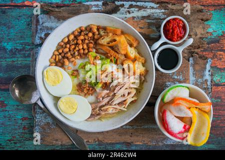 Bubur ayam, porridge di riso indonesiano con pollo sminuzzato e servito con uova sode bollite, cracker di gamberi e sambal di pasta di peperoncino Foto Stock