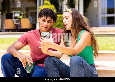 Biracial bella giovane donna che mostra smartphone al ragazzo di caffè mentre si siede sui gradini Foto Stock