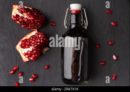 Spremuta di melograno in una bottiglia trasparente di vetro, melograno spezzato e semi di melograno su un piatto di pietra piatta nera Foto Stock