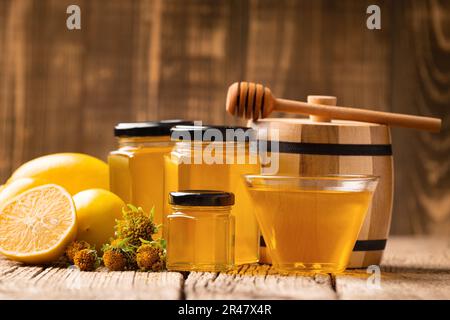 Composizione concettuale di prodotti biologici di api e limoni, miele in vasetti, ciotola trasparente e barile di legno con miele su fondo ligneo. Foto Stock