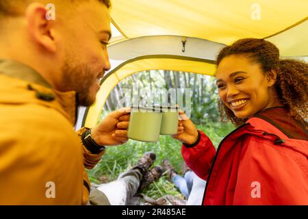 Felice coppia biraciale bere tè e sedersi in tenda nella foresta, inalterato Foto Stock