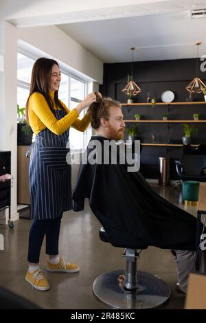 Happy caucasian femmina parrucchiere undoing capelli mordente di cliente maschio a capelli lunghi al salone di parrucchiere Foto Stock